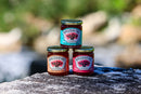 Three jars from the Brushfire Farms Three Amigos collection—featuring bold flavors like Prickly Pear Pepper, Peach Pepper, and Original Pepper—are stacked on a rock outdoors. With green foliage softly blurred in the background, these unique jams from Brushfire Farms make a perfect addition to any gift box or special occasion, especially with a customized logo.