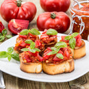 A white plate with toasted bread topped with tomato bruschetta, fresh basil, and SALTOPIA Infused Sea Salts' MYSTICAL PIZZA Tomato & Italian Herbs Seasoning, surrounded by tomatoes and a jar of bruschetta on a wooden table.