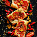 Three rectangular puff pastry tarts topped with tomatoes, yellow chili peppers, herbs, cheese, and SALTOPIA Infused Sea Salts SHE DEVIL Spicy Herbal Blend are surrounded by red chili peppers, herbs, and Pink Himalayan sea salt on a dark surface.