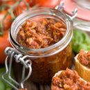 An open glass jar of chunky tomato spread sits on a table, surrounded by fresh tomatoes and basil. Nearby, slices of bread are topped with the spread and finished with SALTOPIA BLT Salt: BACON. LETTUCE. TOMATO. for extra flavor.