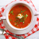 A bowl of tomato soup garnished with cream, herbs, and SALTOPIA Infused Sea Salts' Cheese Salt: CHEDDAR CHEESE sits on a red-and-white checkered napkin, with a spoon beside it.