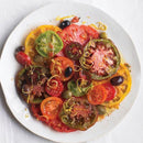 A white plate with colorful heirloom tomato slices, olives, capers, and lemon zest is finished with SALTOPIA Infused Sea Salts' Saltopia BLT Salt: BACON. LETTUCE. TOMATO., served on a white tablecloth.