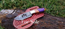 A Texan Knives Traditional Cowboy Knife – 8" resin & olivewood handle, steel bolster, and leather sheath rests on a log with green grass behind it. "For Sale" text is displayed on the image.