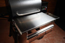 A Pimp My Grill® Tactical Stainless Folding Front Shelf attached to a pellet grill, serving as a stainless steel folding front prep station.