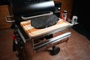 A Pimp My Grill® Tactical Stainless Folding Front Shelf attached to a pellet grill holds a wooden cutting board with a smoked brisket, spice rub, and beverage, with butcher paper stored on a rack below.