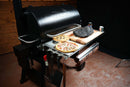 A Pimp My Grill® Tactical Stainless Folding Front Shelf attached to a Traeger Pro 780, holding a pizza, a pie, and a wooden cutting board with a smoked brisket.