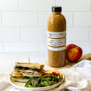A quart bottle of Terrapin Ridge Farms Apple Cider Maple Balsamic Vinaigrette stands by a peach. A sandwich on multigrain bread, apple slices, and mixed greens are arranged on a white plate with a light napkin in front.