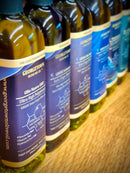 Several bottles of Georgetown Olive Oil Co. 6 Bottle Variety Pack, adorned with blue and gold accents, are arranged in a row on a wooden surface. The front bottle’s label features "Ultra High Polyphenols Extra Virgin Olive Oil" from Georgetown Olive Oil Co., offering the rich flavors of Greek extra virgin olive oil and the essence of Mediterranean taste.