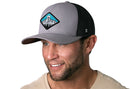A smiling man with short brown hair and a trimmed beard wears the Haka Hat Diamond Mountain and Trees Trucker Hat, a gray-black outdoors snapback by Haka Hat. He is dressed in a light beige shirt against a white background.