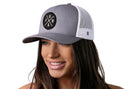 A smiling woman wears the Haka Hat PNW Trucker Hat—Gray-White PNW X Snapback, featuring a circular black patch with "P", "N", "W", and an arrow symbol, plus a comfort-fit sweatband for all-day wear.