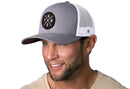 Smiling man with light facial hair wears a Haka Hat PNW Trucker Hat, Gray-White PNW X Snapback featuring a black round patch with crossed arrows and P,N,W letters. He pairs it with a light beige shirt. Comfort-fit sweatband for all-day ease.
