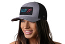 A woman with long brown hair smiles while wearing the Haka Hat Send It Trucker Hat in gray-black, a snapback from Haka Hat featuring a colorful "Send It" patch and comfort-fit sweatband, against a white background.