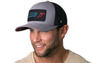 A man with light stubble wears the Haka Hat Send It Trucker Hat in gray-black, featuring a colorful SEND IT patch. The comfort-fit sweatband from Haka Hat ensures all-day ease. He is dressed in beige and facing slightly left.