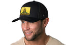 A smiling man with a trimmed beard wears the Haka Hat Don't Tread On Me Trucker Hat, a black snapback featuring a yellow Gadsden Flag patch with coiled snake. He pairs it with a light shirt against a white background.