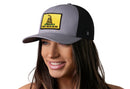 A smiling woman wears the Haka Hat Don't Tread On Me Trucker Hat, a gray-black snapback with a yellow Gadsden Flag patch. This bold hat by Haka Hat completes any look. The background is white.