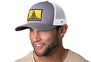 A smiling man with light facial hair wears a Haka Hat Dont Tread On Me Trucker Hat, a gray-white snapback featuring the yellow Gadsden flag with a coiled snake. He is dressed in a beige shirt.