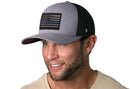 A smiling man with short hair and a trimmed beard wears a Haka Hat Thin Blue Line Trucker Hat, a gray-black police tactical snapback, paired with a beige shirt.