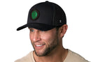 A smiling man wears the Haka Hat Hops Trucker Hat, a black beer snapback by Haka Hat featuring a green hop emblem, paired with a light shirt against a plain white background.