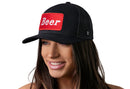A woman with long brown hair smiles while wearing the Haka Hat Beer Trucker Hat, a black drinking snapback by Haka Hat, featuring a red "Beer" patch in white letters. The premium hat pops against the clean white background.