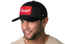 A man wearing the Haka Hat Beer Trucker Hat, a black mesh drinking snapback with a red patch labeled "Beer" in white letters, smiles and looks slightly to the side.