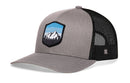 The Haka Hat Mountains and Sky Trucker Hat features a gray-black design, mesh back, and front patch with mountain scene and blue sky. This Haka Hat snapback blends style with outdoor vibes.
