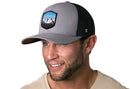 A smiling man with a short beard wears the Haka Hat Mountains and Sky Trucker Hat—gray-black outdoors snapback by Haka Hat—and a beige T-shirt, posing against a white background.