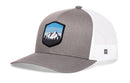 The Haka Hat Mountains and Sky Trucker Hat is a gray-white outdoors snapback with a mesh back and front patch showing mountains, trees, and a blue sky.