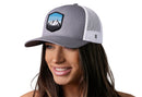 A woman in a gray-white Haka Hat Mountains and Sky Trucker Hat with a mountain and sky patch on the front is smiling and looking slightly to the side.