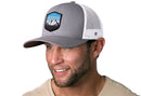 A smiling man with short brown hair and a trimmed beard wears the Haka Hat Mountains and Sky Trucker Hat—gray-white with a mountain patch—paired with a beige shirt. The background is white.