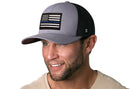 A smiling man with a short beard wears a Haka Hat Thin Blue Line Trucker Hat, Gray-Black Police Snapback, featuring a black and white American flag with a blue stripe. He has a beige shirt and stands against a white background.