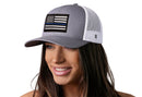 A smiling woman wears a Haka Hat Thin Blue Line Trucker Hat—gray and white with a black, white, and blue American flag patch—by Haka Hat. The background is plain white.