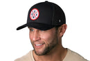 A smiling man with stubble wears the Haka Hat USA Trucker Hat in Black America Snapback, featuring a mesh back and circular embroidered patch. He pairs it with a light beige shirt against a white background.