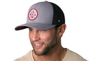 A smiling man with a short beard and mustache wears the Haka Hat USA Trucker Hat in gray-black, featuring a circular USA logo. He has light skin, a beige shirt, and stands against a white background. Brand: Haka Hat.