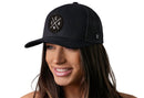 A woman with long brown hair smiles, wearing the Haka Hat USA Trucker Hat | Black America Snapback by Haka Hat, featuring a black mesh design and an embroidered X patch with "U S A" on the front. The background is plain white.