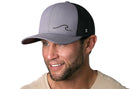 A smiling man with a trimmed beard wears the Haka Hat Wave Trucker Hat—gray-black beach snapback with a minimalist wave logo—paired with a beige shirt, set against a white background.