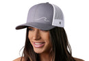 A smiling woman wears the Haka Hat Wave Trucker Hat | Gray-White Beach Snapback by Haka Hat, featuring a white wave design on the front, shown against a plain white background.