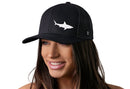 A woman with long brown hair smiles while wearing the Haka Hat Shark Trucker Hat, a black beach snapback by Haka Hat featuring a white shark design on the front. The background is plain white.