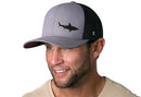 A smiling man with a short brown beard wears the Haka Hat Shark Trucker Hat in gray and black, featuring a shark silhouette. He pairs it with a light beige t-shirt against a white background. Brand: Haka Hat.
