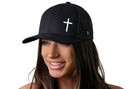 A woman with long brown hair smiles while wearing the Haka Hat Cross Trucker Hat, a black snapback by Haka Hat featuring a white cross on the front. The background is plain white.
