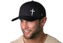 A man with a trimmed beard wears a beige shirt and the Haka Hat Cross Trucker Hat in Black, smiling and looking slightly to the side against a white background.