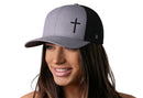 A woman with long brown hair smiles wearing the Haka Hat Cross Trucker Hat, a gray-black snapback by Haka Hat featuring a bold black cross on the front panel, set against a plain white background.