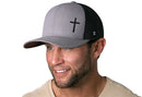 A smiling man with a short beard wears a Haka Hat Cross Trucker Hat—this gray and black snapback from Haka Hat features a bold black cross. He pairs it with a light-colored shirt.