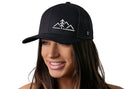 A woman with long brown hair smiles while wearing the Haka Hat Mountains with Pine Tree Trucker Hat, a black outdoors snapback by Haka Hat, featuring a white mountain and tree logo. The background is plain white.