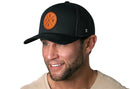 A man with a trimmed beard smiles while wearing the Haka Hat PNW Trucker Hat—an all-black snapback by Haka Hat featuring a circular brown leather patch with "PNW"—paired with a beige shirt, set against a white background.
