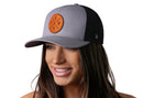 A smiling woman wears a Haka Hat PNW Trucker Hat in gray and black with a brown leather patch snapback featuring crossed "PNW" letters on the front. The background is white.