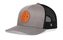 The Haka Hat PNW X Snapback is a gray and black mesh-back trucker hat with a curved brim, featuring an orange circular patch with crossed lines, the letters N, P, W, and an arrow on the front.