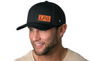 A man in a Haka Hat LFG Trucker Hat | Black Lets Go Snapback, featuring an orange LFG patch and comfort-fit sweatband, smiles slightly to the side. He has light facial hair, a beige T-shirt, and stands against a white background.