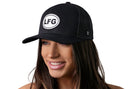 A smiling woman with long brown hair wears the Haka Hat LFG Trucker Hat, a black "Let's Go" snapback by Haka Hat, featuring a white circular "LFG" patch on the front. The hat stands out against the plain white background.
