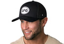 A smiling man with a short beard wears the Haka Hat LFG Trucker Hat in black, featuring a white circular Lets Go logo, paired with a beige shirt against a white background.