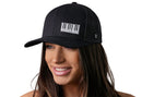 A woman with long brown hair smiles while wearing the Haka Hat Piano Keys Trucker Hat, a black snapback by Haka Hat featuring a white piano keyboard design, standing out against a clean white background.